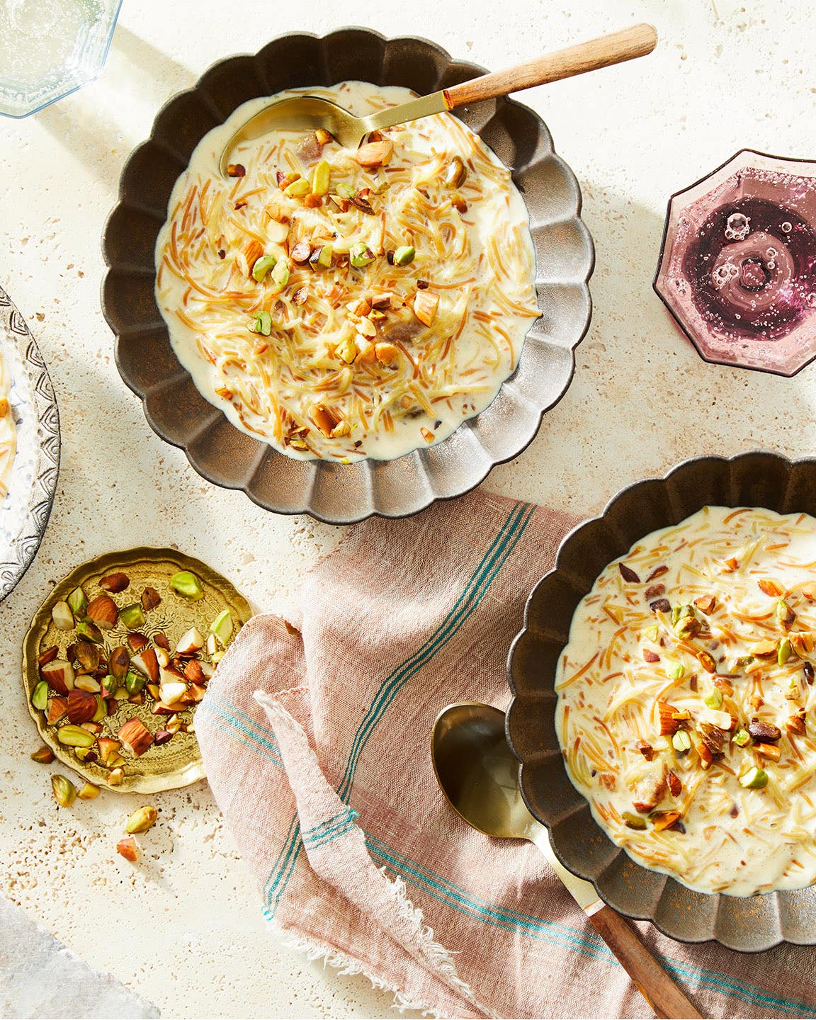 Sheer Khurma Two scalloped bowls of sheer khurma, a creamy milk-and-vermicelli pudding topped with chopped nuts, with spoons alongside on a light tabletop.